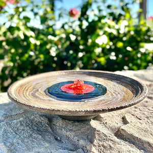 QuackUps Solar Bubbler Fountain on a rock, surrounded by plants, perfect for attracting hummingbirds. Includes solar panel, pump, and red slotted dome.