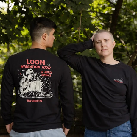 Two people wearing Loons of North America Sweatshirt, Charcoal, showcasing back graphic of a loon in gear and front logo, amidst leafy trees.