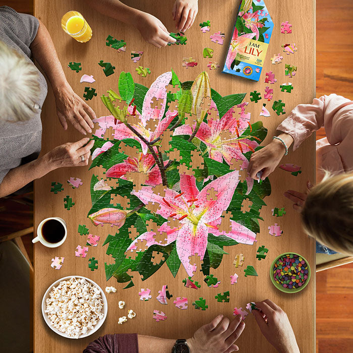 Group assembling the I Am Lily 350 pc. Puzzle, surrounded by snacks and drinks, highlighting its unique floral shape and engaging design.