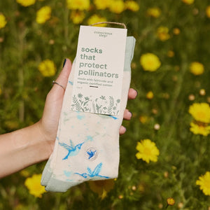 Conscious Step hummingbird socks with gift packaging held in front of yellow garden flowers