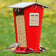 Snack Shack Resistor Feeder: Red, house-shaped bird feeder with clear panels, two perches, filled with seed; titmouse and goldfinch visible, squirrel-resistant design.
