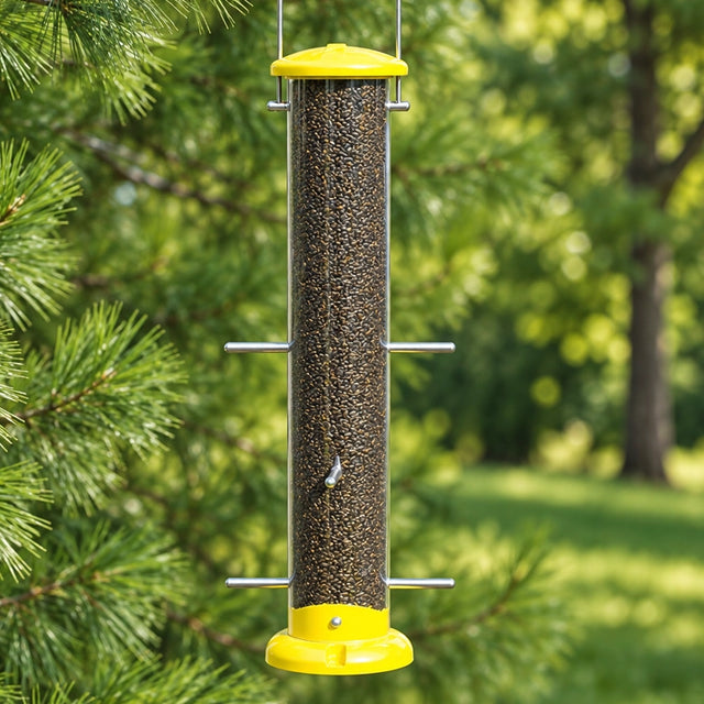 Yellow Nyjer tube bird feeder filled with seed, hanging beside pine branches in a sunlit green backyard.