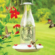 Milkhouse 24 Ounce Glass Hummingbird Feeder, featuring a vintage milk can design, with red flower feeding ports and hummingbirds feeding, against a green backdrop.
