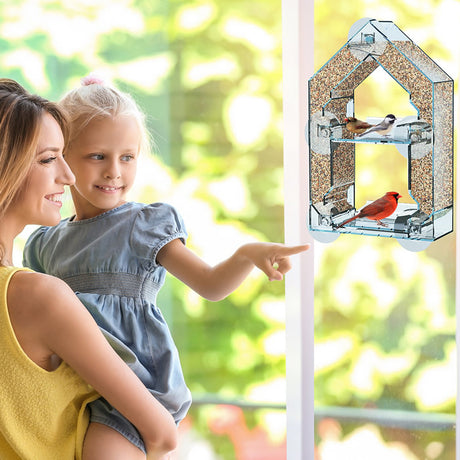 Woman holding a child, observing a Sneak-A-Peek Window Mount Feeder attached to a window, designed for close-up bird watching with a reflective one-way mirror.