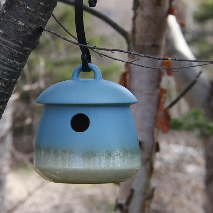 Return Bird Home, Soft Blue, hanging from a tree, featuring a small entrance hole and durable stoneware construction designed for attracting small nesting birds.