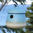 Return Bird Home, Soft Blue, hanging from a tree branch. The stoneware birdhouse features a 1-1/8 inch opening and a durable glaze finish.