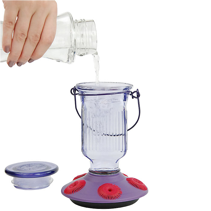 Hand pouring liquid into a blender jar, with a focus on the Lavender Field Top-Fill Glass Hummingbird Feeder featuring zinnia-shaped feeding ports and a see-through glass reservoir.