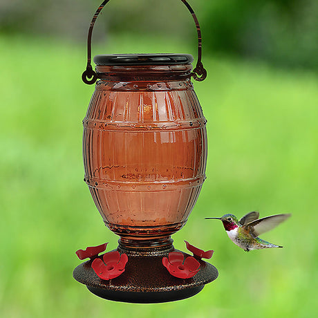 Prohibition Top-Fill Glass Hummingbird Feeder: A hummingbird hovers near a glass jar feeder with flower-shaped feeding ports, inspired by a Prohibition-era barrel design.