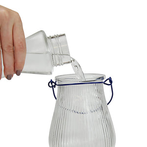 Hand pouring water into the Grand Oasis Top-Fill Glass Hummingbird Feeder with visible clear, striped glass reservoir and blue handle.