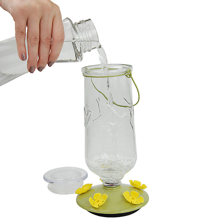 Hand pouring water into a Perky-Pet Desert Bloom Top-Fill Hummingbird Feeder with five feeding ports and a clear glass bottle.