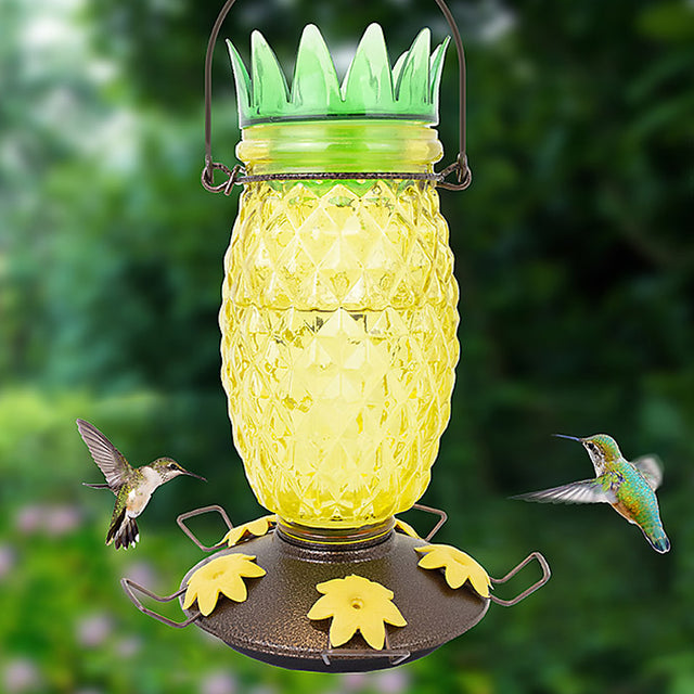 Perky-Pet Pineapple Top-Fill Hummingbird Feeder with hummingbirds flying around, featuring a yellow glass jar, green lid, and multiple feeding ports.