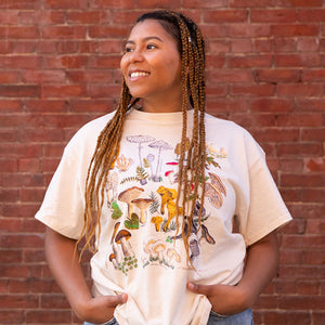 Woman wearing a Mushrooms T-Shirt with a nature scene design by Ieva Tatarsky, smiling with braided hair.